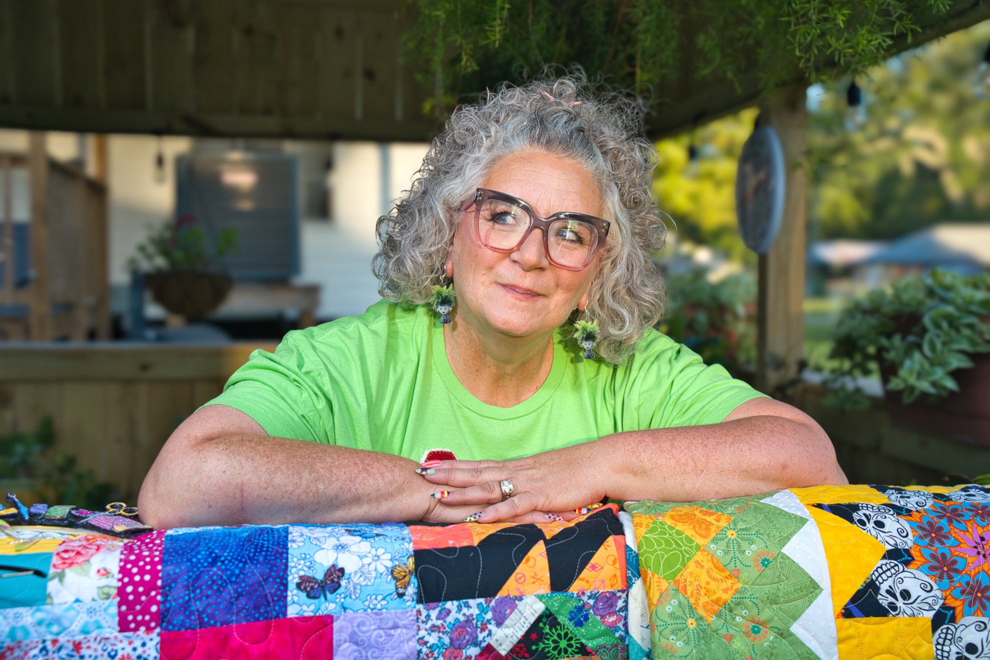Angela in a green tee, arms folded on a colourful patchwork quilt under the garden pergola, smiling at the camera