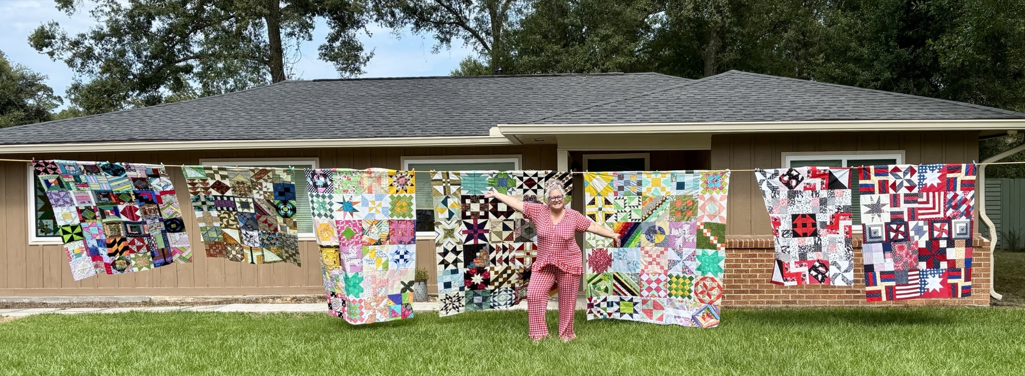 Angela with her arms spread wide in front of seven colourful patchwork quilts hung on a line across her front lawn